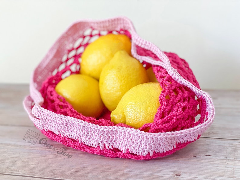 May include: A bright pink and light pink crocheted net bag containing four yellow lemons. The bag features a wide, solid crocheted border at the top and a mesh body. A small logo with the text One & Two is visible on the lower left side.