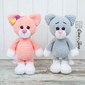 May include: Two crocheted cat plushies, one pink with white accents and a flower on its head, and one grey with white accents. Both have black eyes and pink noses.