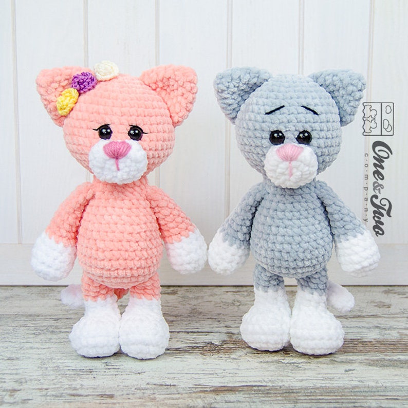 May include: Two crocheted cat plushies, one pink with white accents and a flower on its head, and one grey with white accents. Both have black eyes and pink noses.