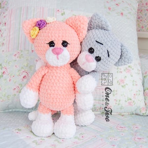 May include: Two crocheted cat plushies, one pink with white accents and a flower crown, the other grey with white accents. Both have black eyes and pink noses.