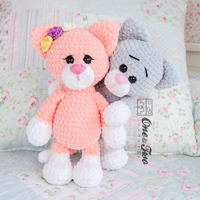 May include: Two crocheted cat plushies, one pink with white accents and a flower crown, the other grey with white accents. Both have black eyes and pink noses.