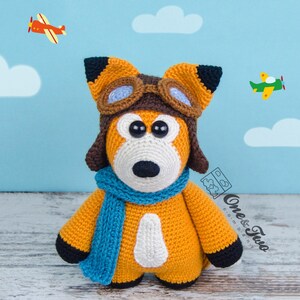 May include: A crocheted orange fox wearing a brown aviator hat with goggles and a blue scarf. The fox is standing on a wooden surface with a blue background with white clouds and two red and green airplanes.