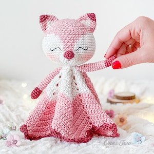 May include: A handmade crochet fox toy in shades of pink and white. The fox has a closed-eye expression, a small pink nose, and a matching pink and white patterned head and ears. The toy has a blanket-like body with a scalloped edge.