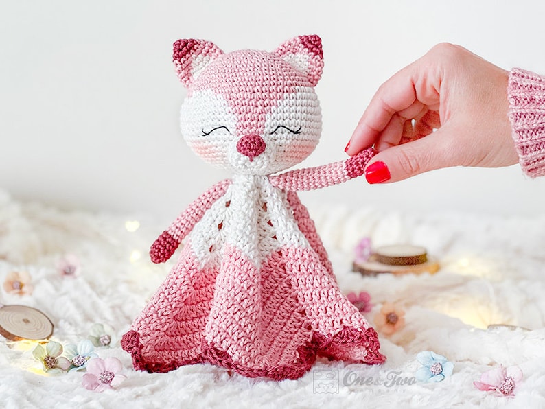 May include: A handmade crochet fox toy in shades of pink and white. The fox has a closed-eye expression, a small pink nose, and a matching pink and white patterned head and ears. The toy has a blanket-like body with a scalloped edge.