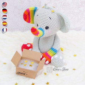 Könnte beinhalten: Ein grauer gehäkelter Elefant mit Regenbogenstreifen an seinen Ohren, seinem Rüssel und seinen Beinen. Der Elefant sitzt auf einer weißen flauschigen Oberfläche mit einer kleinen Pappbox, auf der "Stars" steht. Die Box ist geöffnet und mit goldenen sternförmigen Konfetti gefüllt.