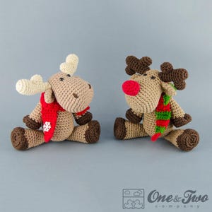Puede incluir: Dos animales de peluche tejidos a crochet, un reno y un alce, ambos con bufandas. El reno tiene la nariz roja y una bufanda verde y roja. El alce tiene una bufanda roja con un copo de nieve blanco.