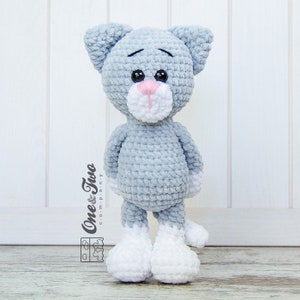 May include: A crocheted grey cat toy with white feet and a pink nose. The cat has a sad expression and is standing on a wooden surface.