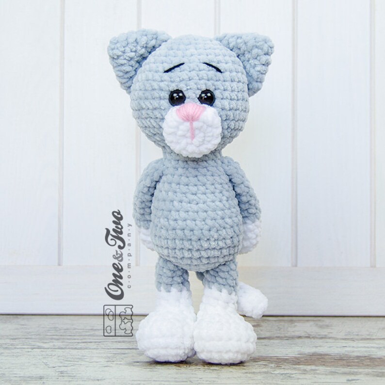 May include: A crocheted grey cat toy with white feet and a pink nose. The cat has a sad expression and is standing on a wooden surface.