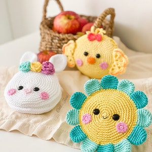 May include: Three crocheted plush items: a white bunny with a floral crown, a yellow chick with a red bow, and a yellow flower with teal petals. Each item features black bead eyes and pink stitched cheeks, crafted with detailed yarn textures.