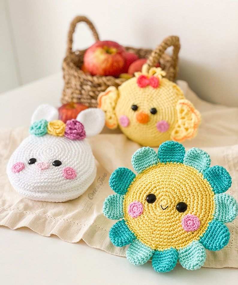 May include: Three crocheted plush items: a white bunny with a floral crown, a yellow chick with a red bow, and a yellow flower with teal petals. Each item features black bead eyes and pink stitched cheeks, crafted with detailed yarn textures.