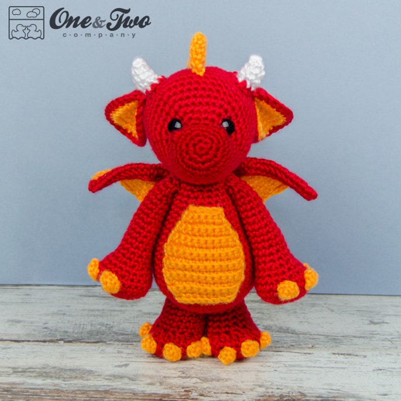 K&ouml;nnte beinhalten: Ein rotes und oranges H&auml;kelspielzeug in Form eines Drachen mit wei&szlig;en H&ouml;rnern. Der Drache hat einen freundlichen Ausdruck und steht auf einer Holzoberfl&auml;che.