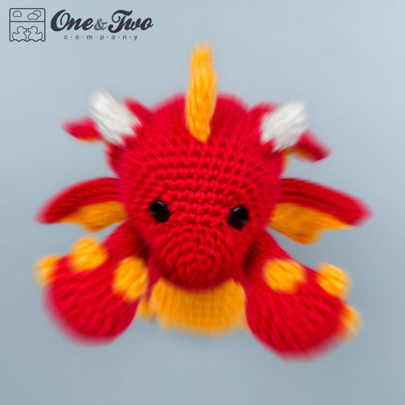 K&ouml;nnte beinhalten: Ein roter H&auml;kel-Drache mit gelben und wei&szlig;en Akzenten. Der Drache hat einen kleinen, runden K&ouml;rper mit einem langen, spitzen Schwanz und zwei kleinen Fl&uuml;geln. Die Augen des Drachen sind schwarz und perlenf&ouml;rmig.