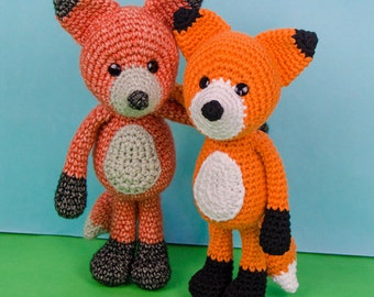 May include: Two crocheted fox plushies, one orange and one peach, with black accents on the ears, paws, and tails. The foxes are standing on a green surface. One & Two Company.