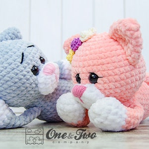May include: Two crocheted plush toys, one grey and one pink, with white accents. Both toys have large eyes and a sweet expression. The grey toy is lying down with its head turned to the side, while the pink toy is lying on its stomach with its head raised. The pink toy has a flower on its head.