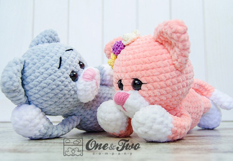 May include: Two crocheted plush toys, one grey and one pink, with white accents. Both toys have large eyes and a sweet expression. The grey toy is lying down with its head turned to the side, while the pink toy is lying on its stomach with its head raised. The pink toy has a flower on its head.