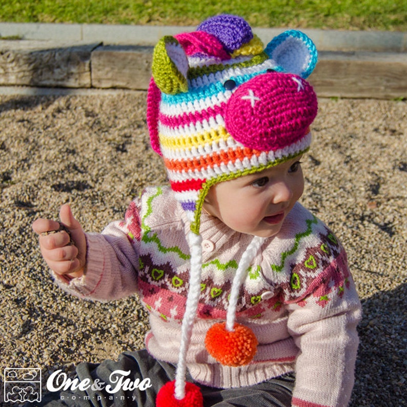 May include: A colorful, handmade crochet hat in the shape of a unicorn's head. The hat features stripes of various colors, including pink, yellow, green, and blue. It has white ear details, a pink muzzle, and a purple horn. The hat is worn by a baby.
