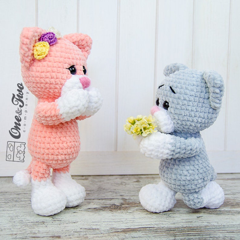 May include: Two crocheted plush toys, a pink cat holding a bouquet of flowers and a grey cat holding a bouquet of yellow flowers. The cat toys are standing on a white wooden surface.