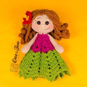 Puede incluir: Una mu&ntilde;eca de crochet con cabello casta&ntilde;o, una blusa rosa y una falda verde. La mu&ntilde;eca lleva una flor en el pelo. La mu&ntilde;eca est&aacute; sentada sobre un fondo amarillo.
