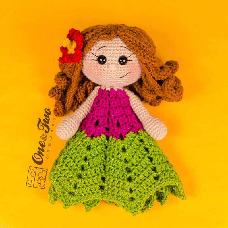 Puede incluir: Una mu&ntilde;eca de crochet con cabello casta&ntilde;o, una blusa rosa y una falda verde. La mu&ntilde;eca lleva una flor en el pelo. La mu&ntilde;eca est&aacute; sentada sobre un fondo amarillo.