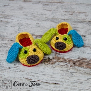 May include: A pair of yellow crocheted baby booties shaped like dogs with blue and green ears. The booties have black eyes, a brown nose, and red trim.