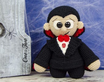 May include: A crocheted vampire doll wearing a black suit with a red bow tie and a cape. The doll has a white face with black eyes and a red mouth.