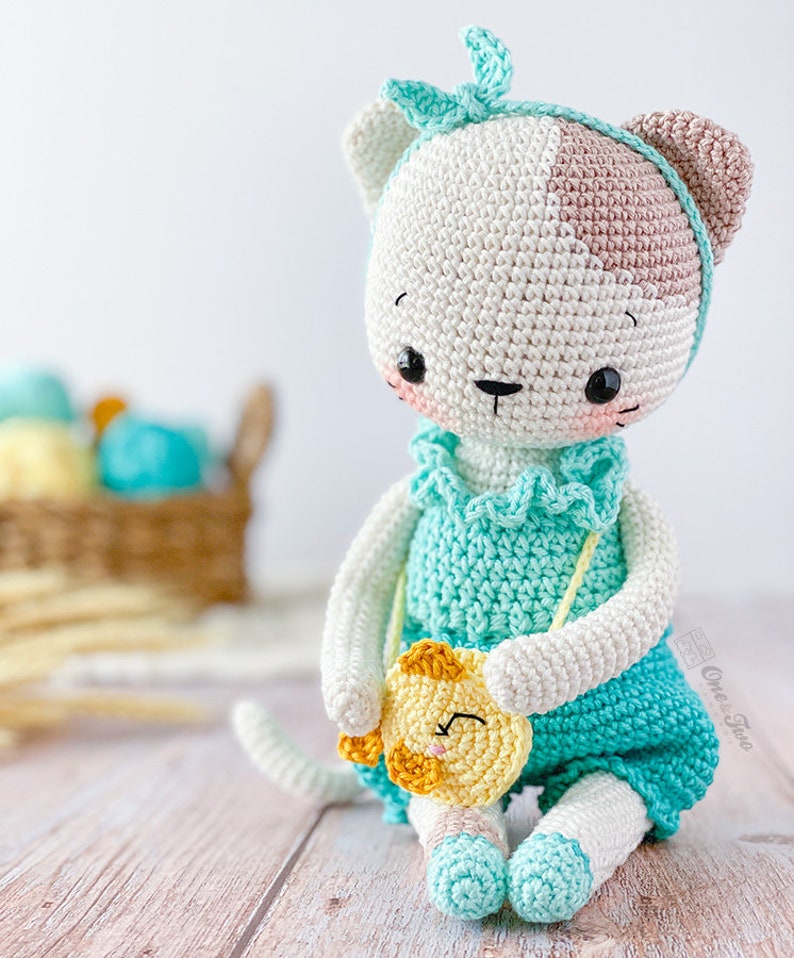 K&ouml;nnte beinhalten: Eine geh&auml;kelte wei&szlig;e Katze mit braunen Ohren, die ein hellblaues Kleid und ein hellblaues Stirnband tr&auml;gt. Die Katze h&auml;lt eine kleine gelbe geh&auml;kelte Katze mit einem l&auml;chelnden Gesicht.