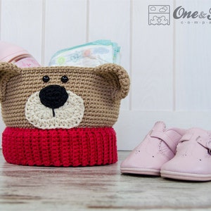 May include: A handmade crochet bear basket in tan, red, and cream. The basket has a bear face design with a black nose and button eyes. A pink baby bottle and diapers are inside. Pink baby shoes are next to the basket.