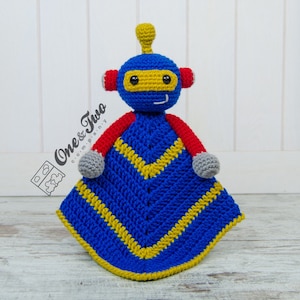 May include: A blue and yellow crocheted baby blanket with a robot attached. The robot has a blue head with yellow eyes and a red body. The blanket has a yellow chevron pattern.