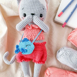 K&ouml;nnte beinhalten: Eine graue geh&auml;kelte Katzendolle, die ein rosa Kleid und Shorts tr&auml;gt und eine blaue geh&auml;kelte Handtasche mit einem Fischdesign h&auml;lt. Die Puppe sitzt auf einer wei&szlig;en Stoffoberfl&auml;che mit Garnkn&auml;uel in Rosa und Grau.