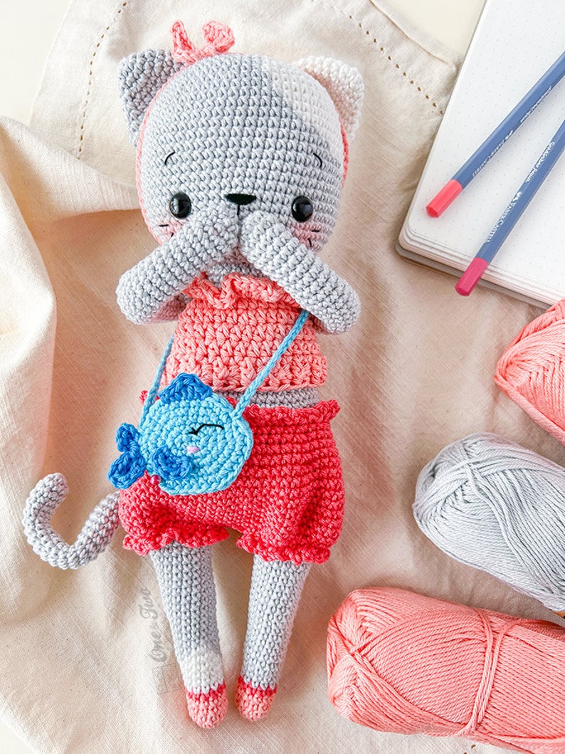 K&ouml;nnte beinhalten: Eine graue geh&auml;kelte Katzendolle, die ein rosa Kleid und Shorts tr&auml;gt und eine blaue geh&auml;kelte Handtasche mit einem Fischdesign h&auml;lt. Die Puppe sitzt auf einer wei&szlig;en Stoffoberfl&auml;che mit Garnkn&auml;uel in Rosa und Grau.