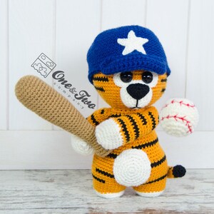 Op de afbeelding: Een gehaakte tijger die een blauwe baseballpet draagt met een witte ster, houdt een bruine honkbalknuppel en een witte honkbal met rode stiksels vast.