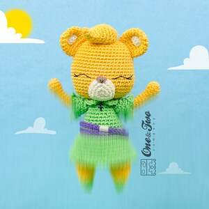 May include: A crocheted yellow bear wearing a green dress with a purple belt. The bear has a white nose and closed eyes. The bear is floating in the air against a blue background with white clouds.