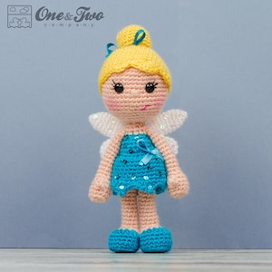 May include: A crocheted fairy doll with blonde hair, a blue dress with sequins, and white wings. The doll is standing on a light blue surface.