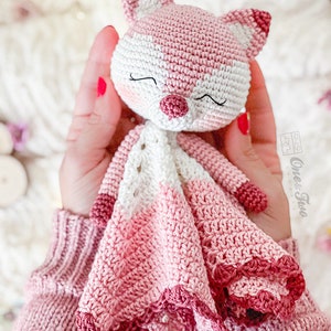 May include: A handmade crochet fox doll in shades of pink and white. The fox has a sweet face with closed eyes and a small, textured nose. The doll's body features a detailed, scalloped edge. The doll is soft and cuddly, perfect for a child.