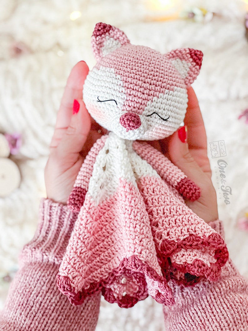 May include: A handmade crochet fox doll in shades of pink and white. The fox has a sweet face with closed eyes and a small, textured nose. The doll's body features a detailed, scalloped edge. The doll is soft and cuddly, perfect for a child.