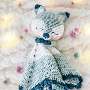 May include: A handmade crochet security blanket featuring a sleeping fox face. The fox is primarily white and gray, with dark blue accents on the ears and nose. The blanket has a scalloped edge in shades of blue and white. The blanket is soft and cozy.