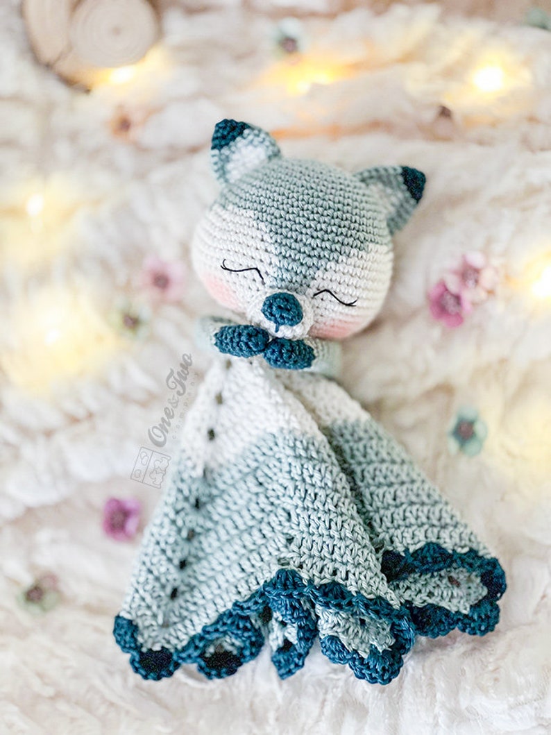 May include: A handmade crochet security blanket featuring a sleeping fox face. The fox is primarily white and gray, with dark blue accents on the ears and nose. The blanket has a scalloped edge in shades of blue and white. The blanket is soft and cozy.