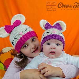 May include: Two children wearing crocheted mouse hats. The older child is wearing a pink and white hat with a pink bow. The younger child is wearing a purple and white hat with a purple bow. The children are smiling and looking at the camera.