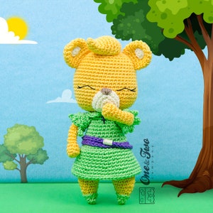 May include: A crocheted plush toy of a yellow bear wearing a green dress with a purple belt. The bear has a white nose and closed eyes. The bear is standing on a green grassy surface with a blue sky and clouds in the background.