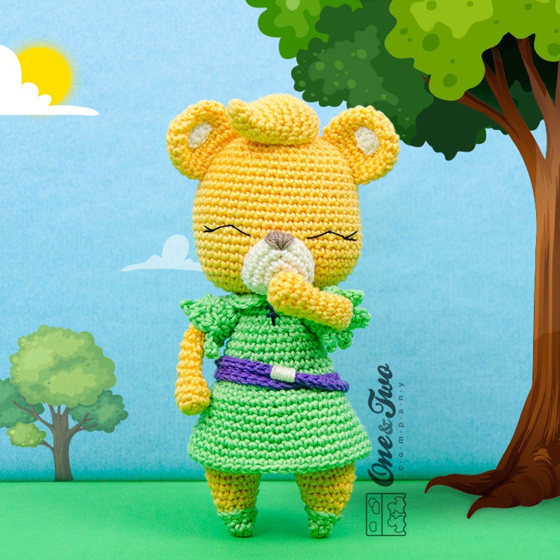 May include: A crocheted plush toy of a yellow bear wearing a green dress with a purple belt. The bear has a white nose and closed eyes. The bear is standing on a green grassy surface with a blue sky and clouds in the background.