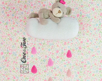 May include: A crocheted mobile with a sleeping bear on a cloud and pink raindrops hanging from it. The mobile is hanging against a floral background.