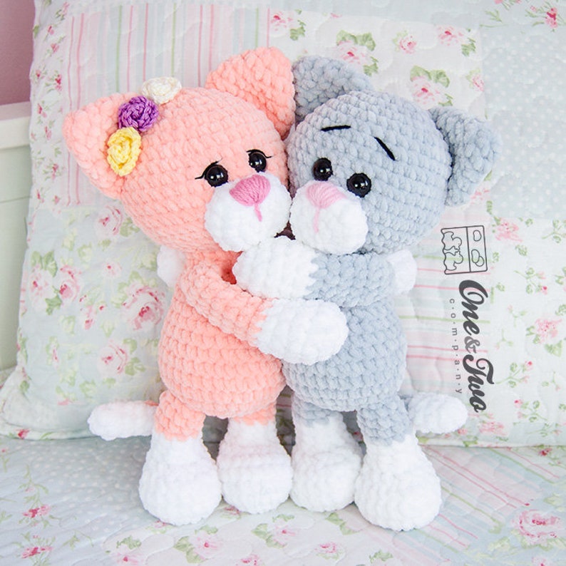 May include: Two crocheted plush cat toys hugging each other. The cat on the left is pink with white paws and a flower crown. The cat on the right is grey with white paws.