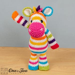 May include: A colorful crocheted zebra toy with rainbow stripes. The zebra has a pink nose and white eyes. The toy is standing on a wooden surface.