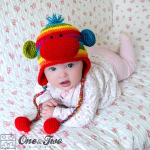 May include: A baby wearing a colorful crocheted monkey hat with a red nose and long, rainbow-colored ties. The hat has green ears and a pom-pom on top.