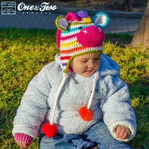 May include: A colorful, crocheted unicorn hat with a pink muzzle, blue ears, and a rainbow mane. The hat has white braided ties with red pom-poms. The hat is worn by a child wearing a blue fleece jacket and jeans.