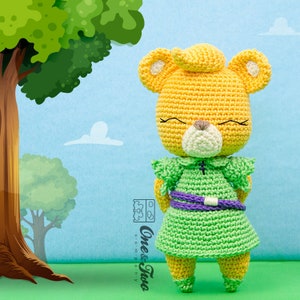 May include: A crocheted yellow bear wearing a green dress with a purple belt. The bear has a white face with a brown nose and closed eyes. The bear is standing on a green surface.