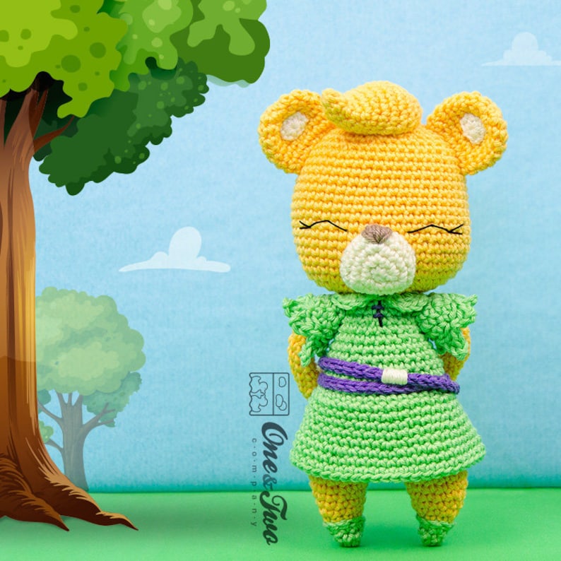 May include: A crocheted yellow bear wearing a green dress with a purple belt. The bear has a white face with a brown nose and closed eyes. The bear is standing on a green surface.
