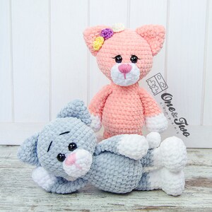 May include: Two crocheted cat toys, one pink with a flower on its head and one grey, lying on a white wood background. The text "One & Two" is visible in the image.