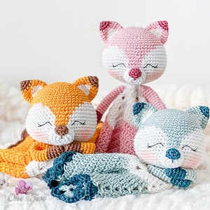 May include: Three handmade crochet animal dolls: a fox in orange and white, a pink fox, and a blue and white bear. Each doll has closed eyes, suggesting sleep. The dolls are on a soft, white surface, possibly a blanket.