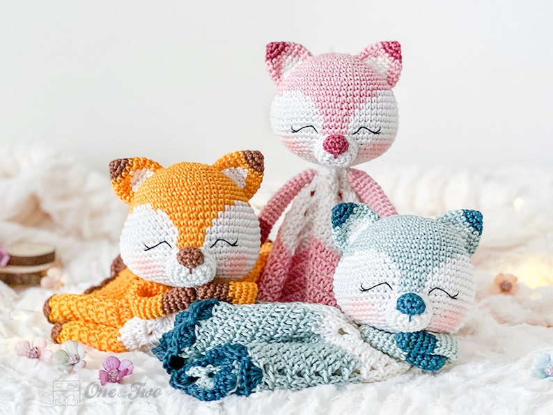 May include: Three handmade crochet animal dolls: a fox in orange and white, a pink fox, and a blue and white bear. Each doll has closed eyes, suggesting sleep. The dolls are on a soft, white surface, possibly a blanket.